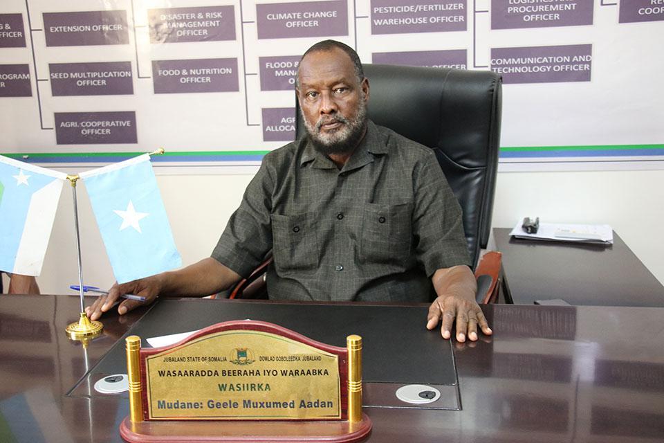 Harmaaruutupaitainen Jubalandin maatalous- ja kasteluministeri Geele Mohamed Adan istuu mustassa nahkatuolissa tumman työpöydän ääressä. Työpöydällä on kullanvärinen nimikyltti sekä Somalian ja Jubalandin osavaltion liput.