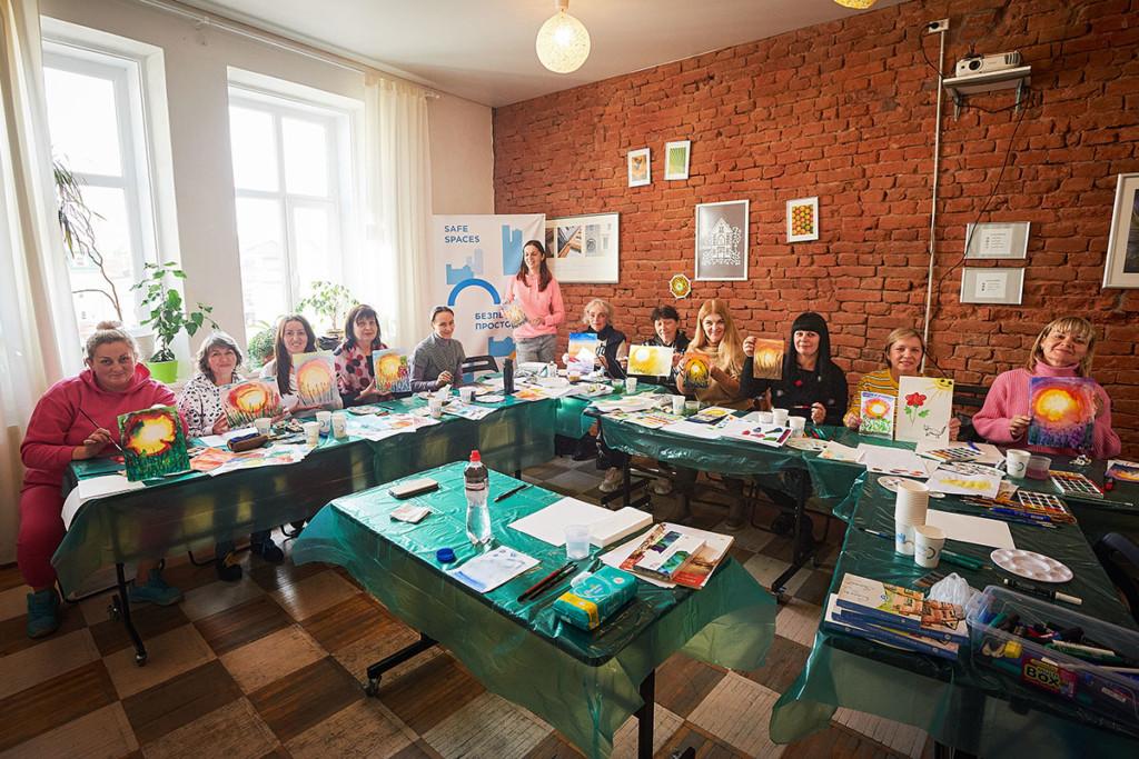 Joukko ukrainalaisia naisia istuu pöytien takana, kaikilla on käsissään itsetehty maalaus. Pöydillä on maalaustarvikkeita, pöydät on suojattu muovilla.