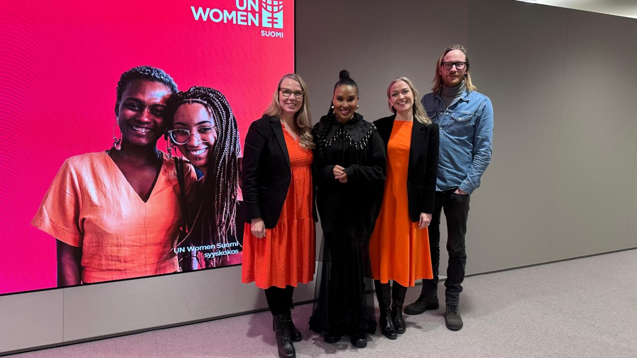 Kuvassa seisoo kolme naista ja yksi mies. Naisilla on päällään oranssia ja mustaa.