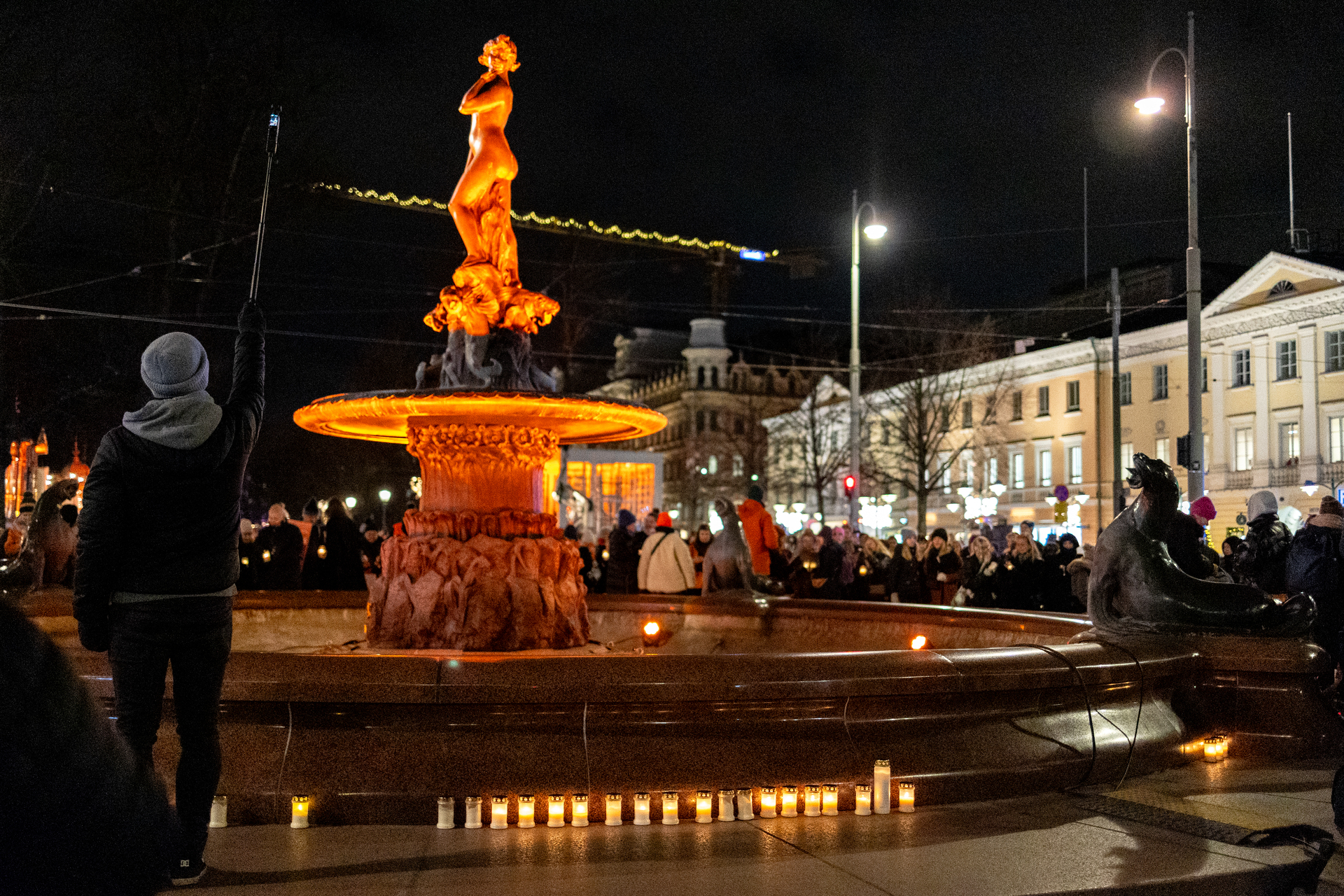 Havis Amanda patsas valaistuna oranssiksi. Patsaan ympärillä on kynttilöitä ja ihmisiä. Kuva: Helsingin kaupunki / Paavo Jantunen